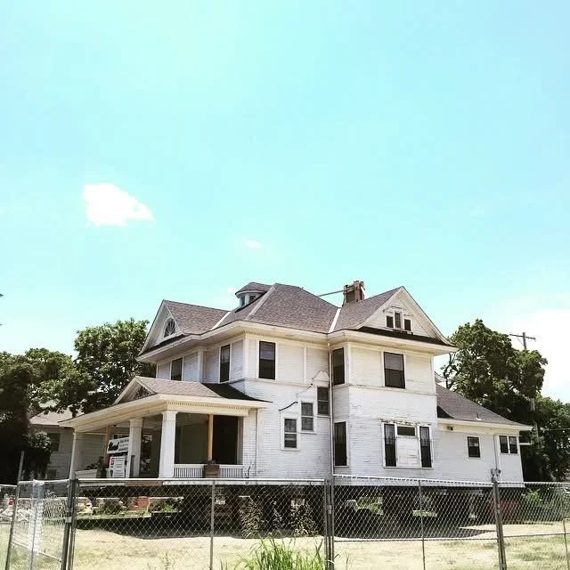 A large white Victorian-style house with multiple gabled roofs, a front porch, and several windows, surrounded by a chain-link fence and trees in the background.