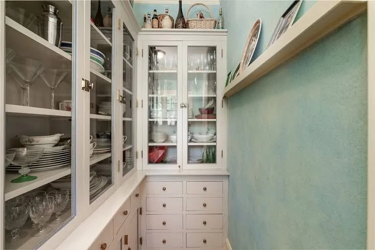 A narrow kitchen cabinet with glass doors filled with dishes, glasses, and bowls, next to a light blue wall with a white shelf holding decorative plates.