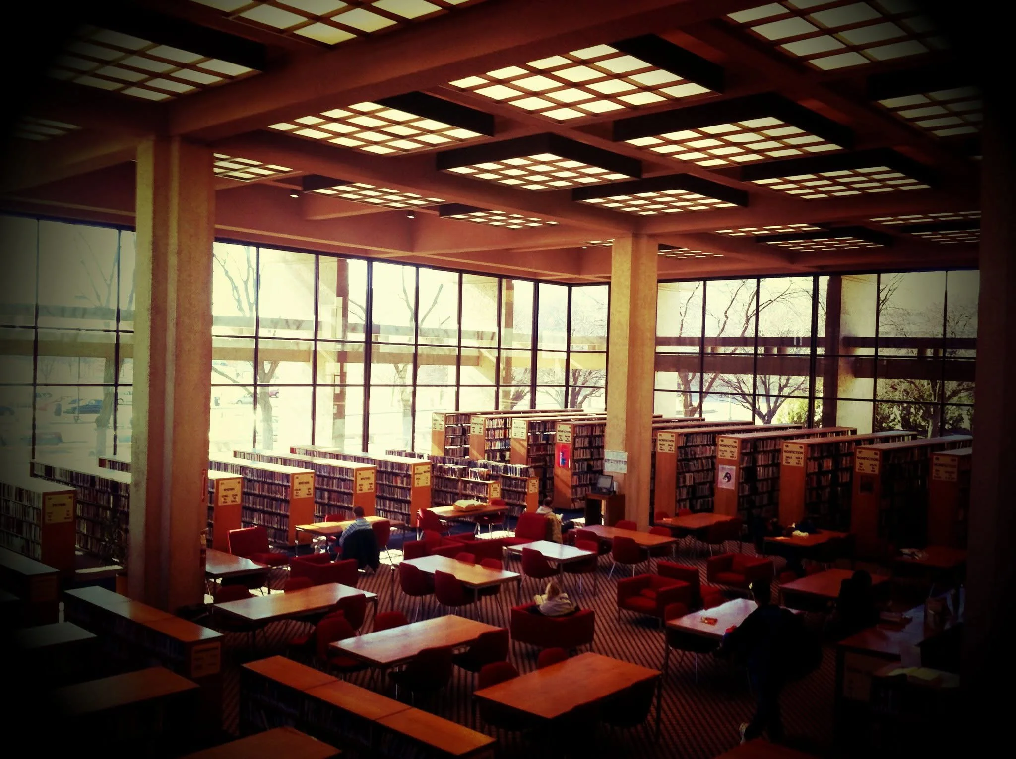 A spacious library with large windows allowing natural light, wooden bookshelves filled with books, red armchairs and tables, and people reading and studying.