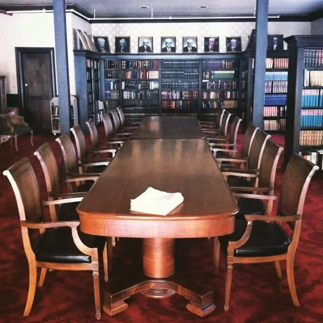 A conference room with a long wooden table surrounded by multiple wooden chairs with black seats. In the background, there are bookshelves filled with books and framed photographs or portraits on top of the shelves.