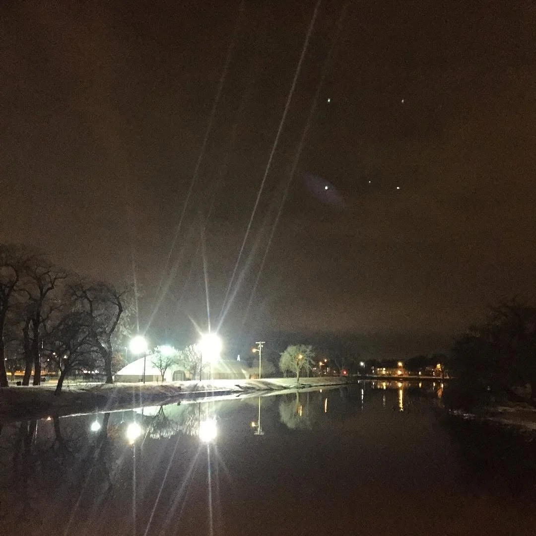 Nighttime scene with a calm river reflecting bright streetlights and bare trees along the shore, under a dark sky with several small and bright stars.