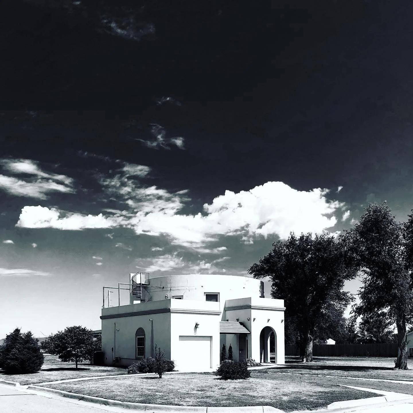 A white, two-story, modern building with a flat roof and arched entrance, surrounded by a well-maintained lawn with small bushes and large trees, under a partly cloudy sky.