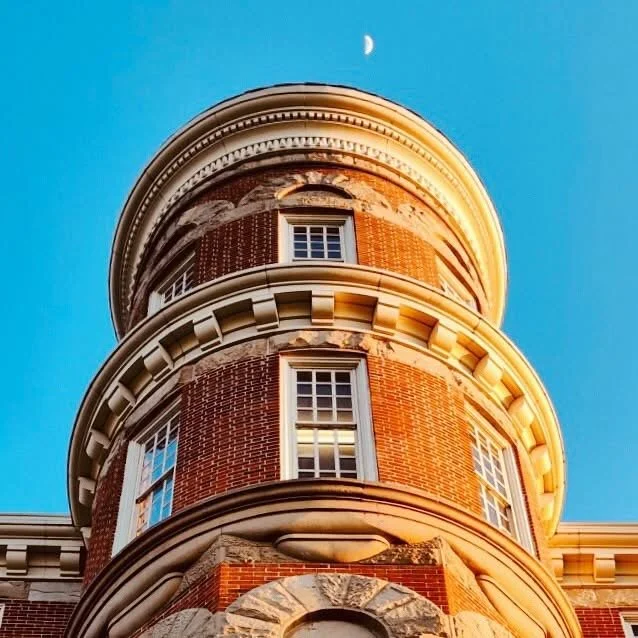 A low-angle view of a historic red brick building with round turrets, white-framed windows, a cornice, and a blue sky with a crescent moon above.
