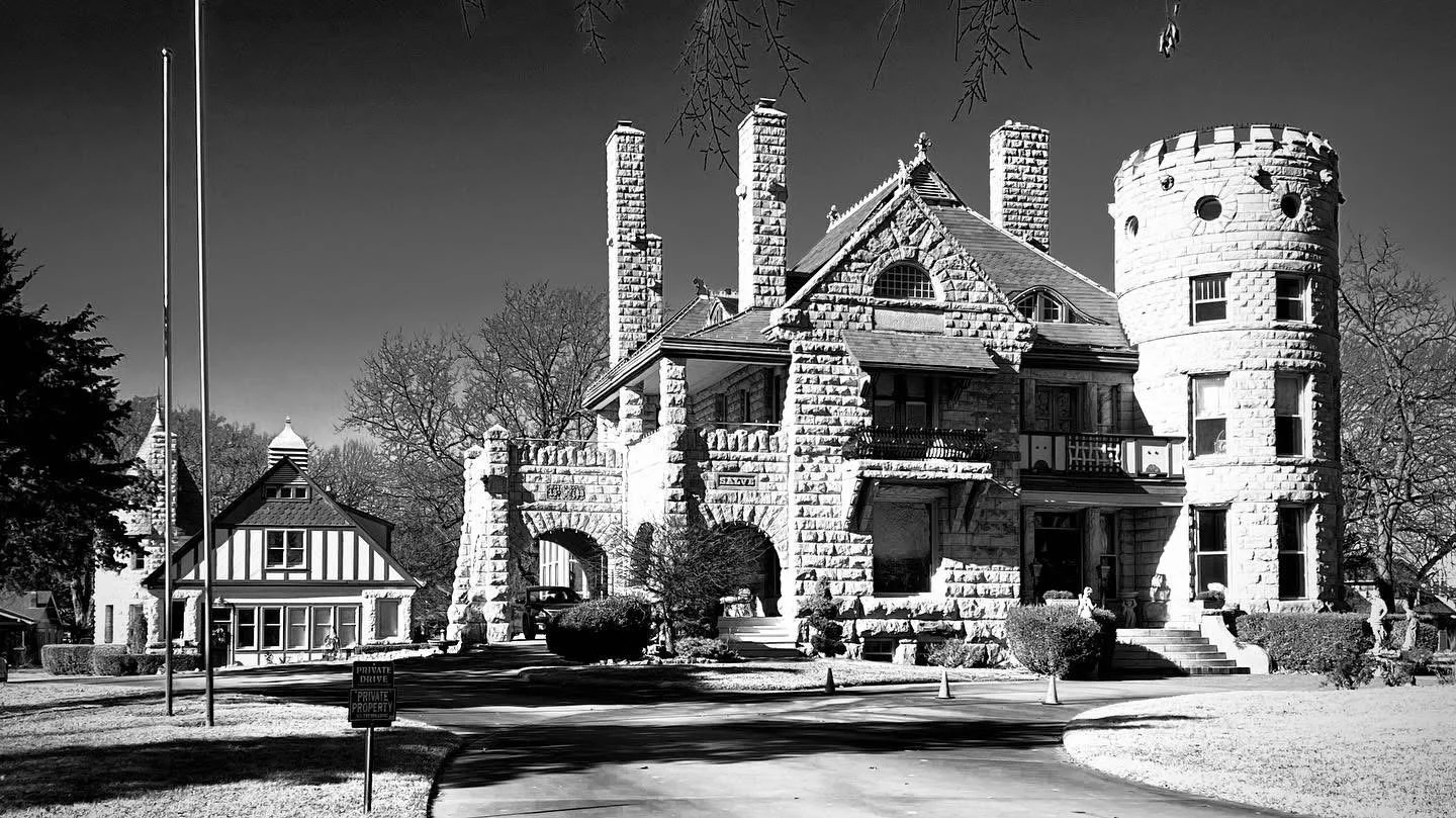 A large stone mansion with a turret, multiple chimneys, and arched entryways, surrounded by trees and a driveway, in black and white.