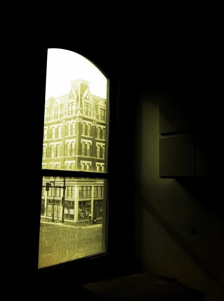 View of a city street through a large window with an arched top, showing a historic building with ornate architecture and a corner storefront.