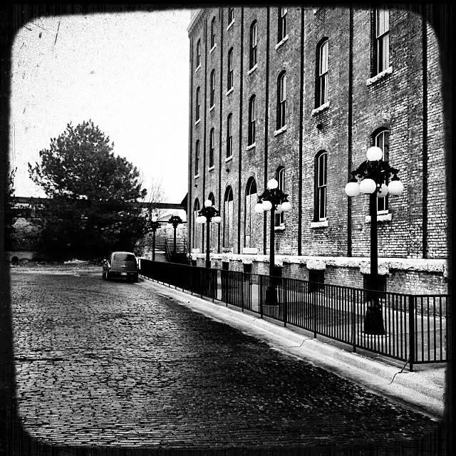 Black and white photograph of a brick building with arched windows, street lamps along the sidewalk, a parked car, and a cobblestone street.