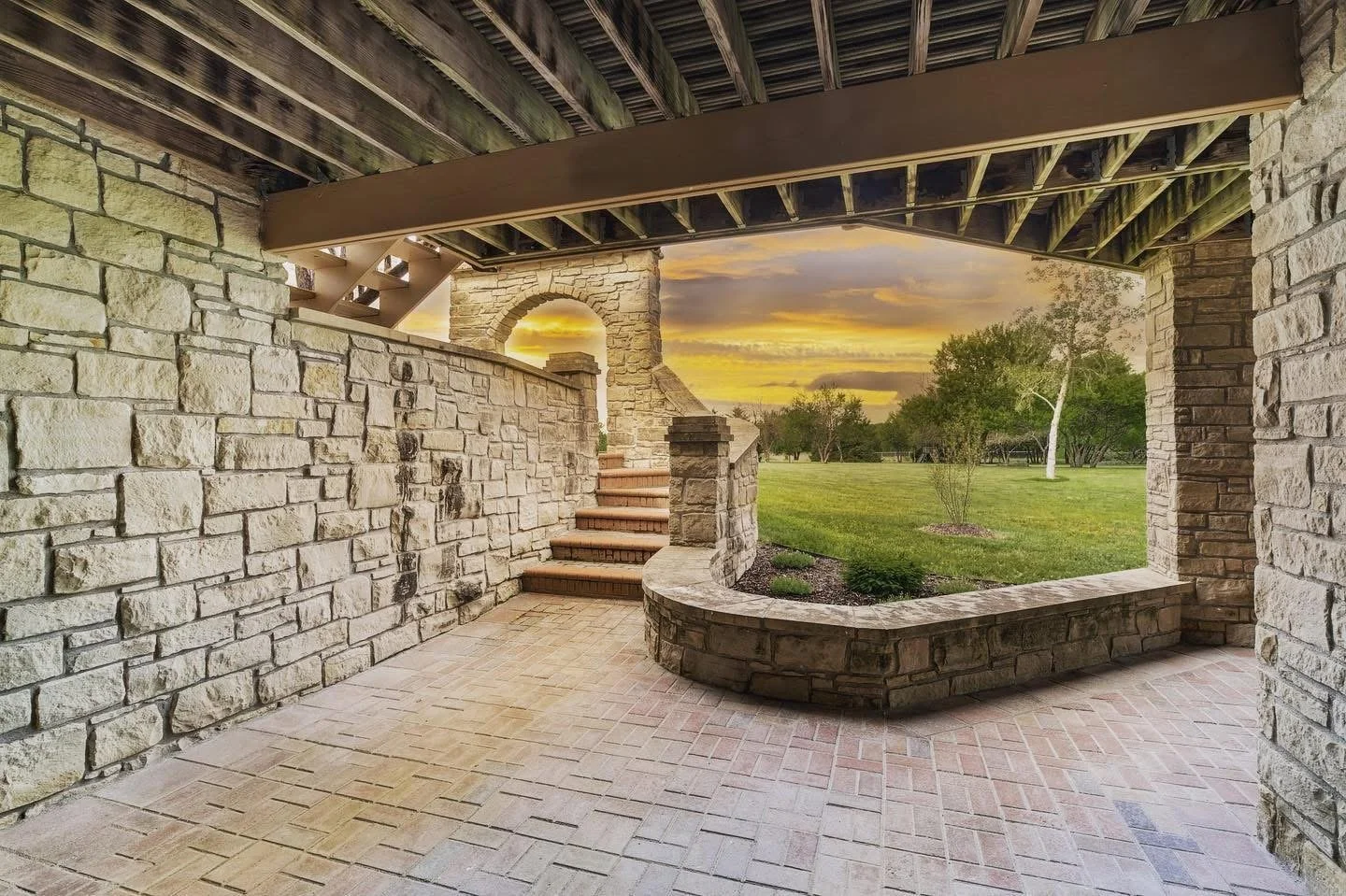 Stone patio with steps leading up to an archway, overlooking a grassy field with trees during sunset.