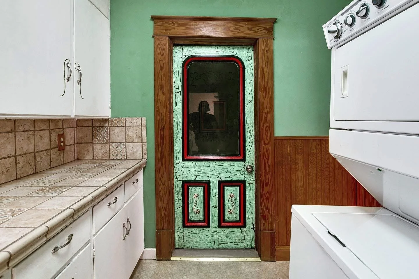 A small kitchen with white cabinets, a tile countertop, and a green painted door with a glass window and decorative panels. The wall is partially paneled with wood, and there is a white washing machine on the right side.