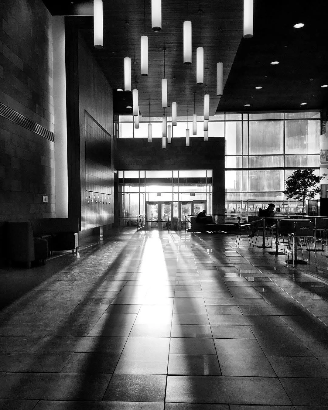 Interior of a modern building lobby with hanging lights, large windows allowing sunlight to cast shadows on the tiled floor, and a few seated people near the windows.