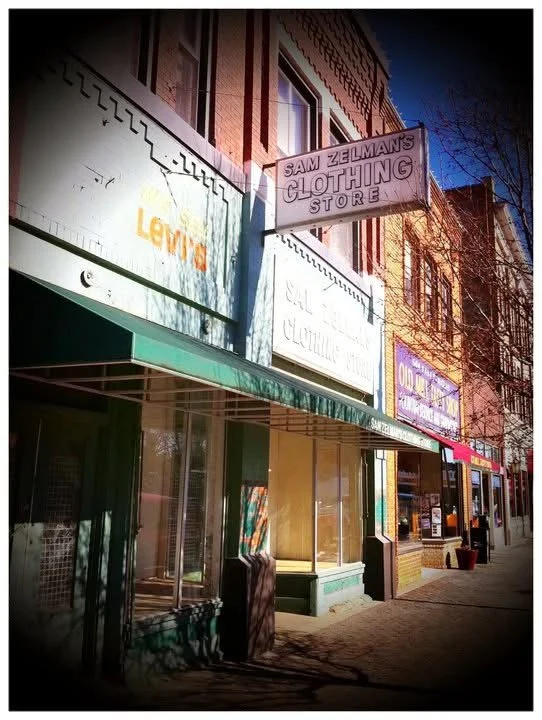 Street view of storefronts, including Sam Zelman's Clothing Store with signs for Levi's and other brands, under a green awning on a sunny day.