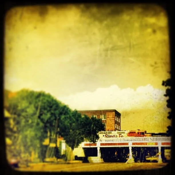 A vintage-style photo of a street scene showing a building, trees, and a Santa Fe train station or train, with a cloudy sky in the background.