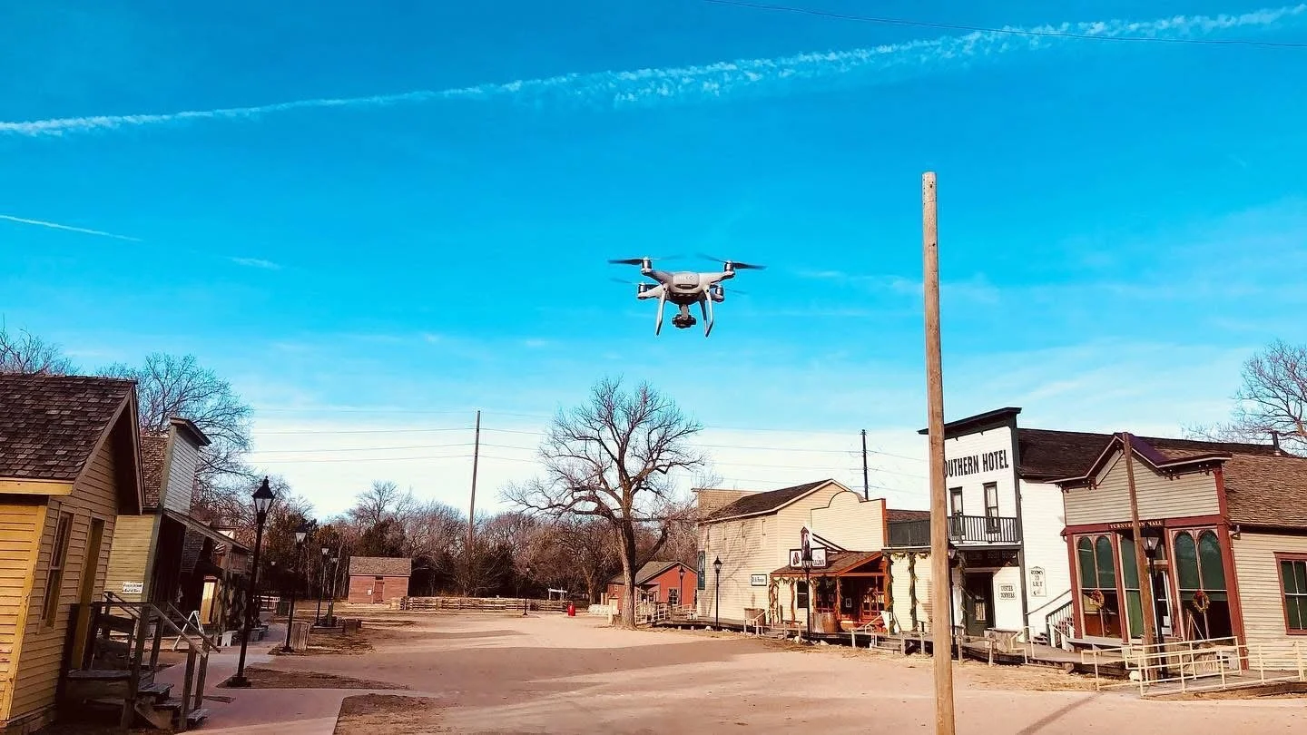 Drone flying over a historic town square with old Western-style buildings and a clear blue sky.