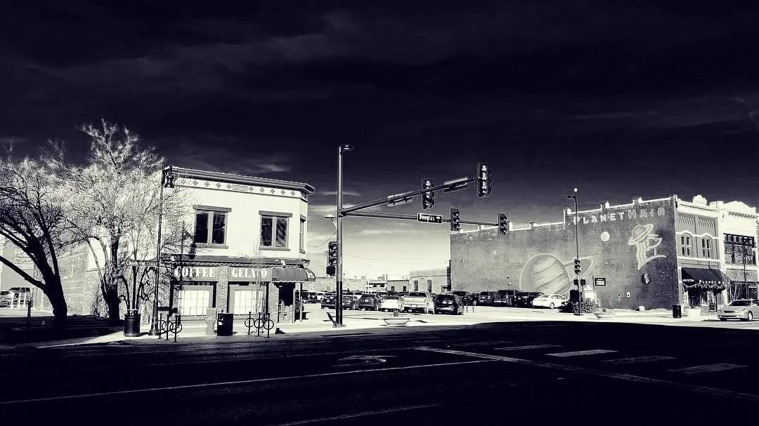 Black and white photo of a street corner with a small building labeled 'Coffee Gelato' on the left and a large building with space-themed mural on the right. Cars are parked along the street and traffic lights are visible.