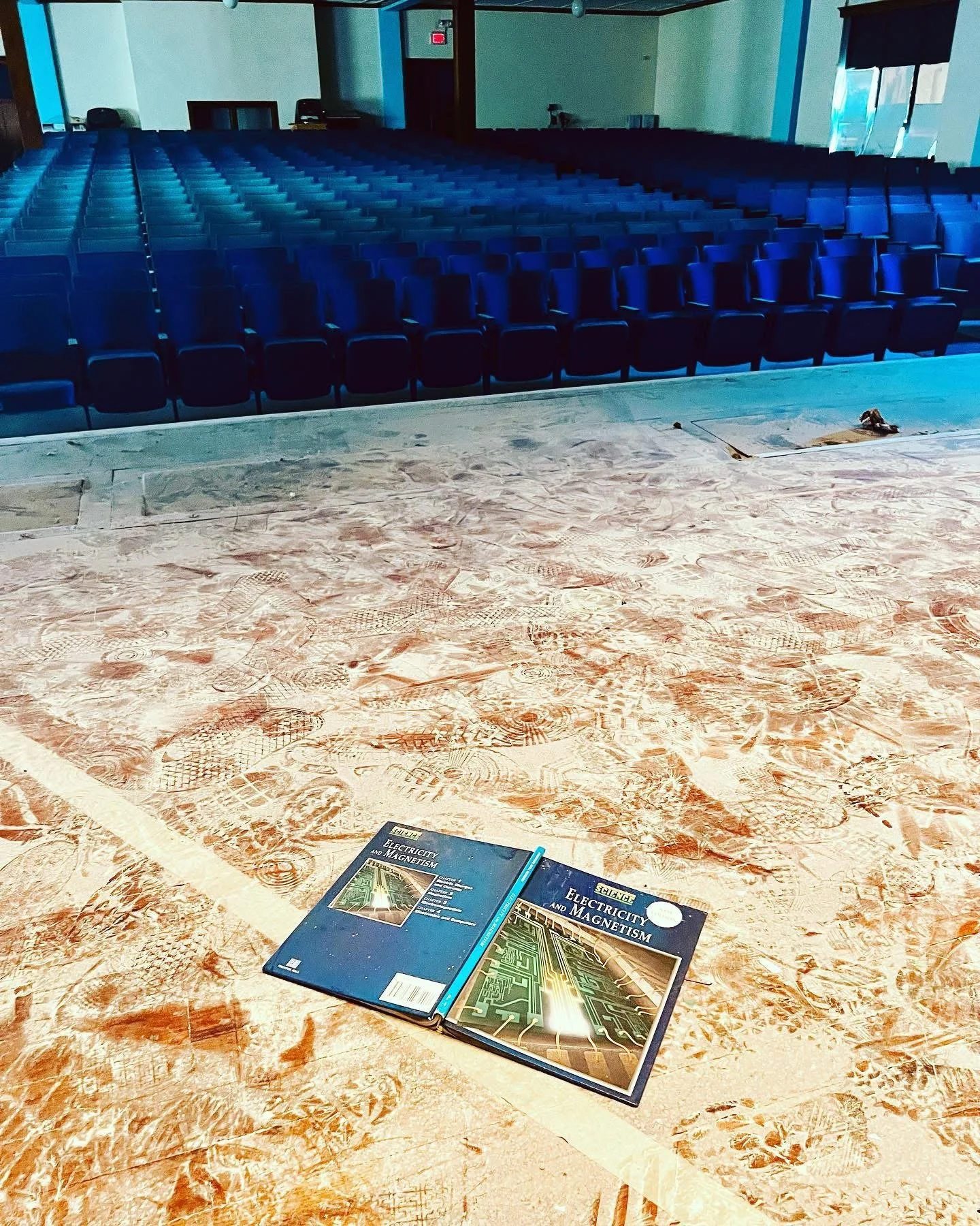 An empty auditorium with blue seats, a stage area with two textbooks titled 'Electricity and Magnetism,' and footprints in the dust on the floor.