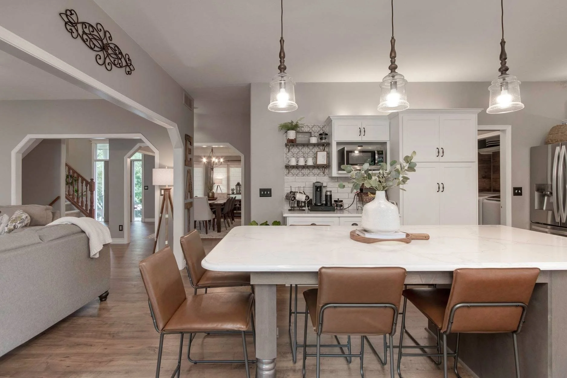 Open concept kitchen with a large white island, brown chairs, gray cabinets, and a coffee station, connected to a dining and living area with wooden floors and modern decor.