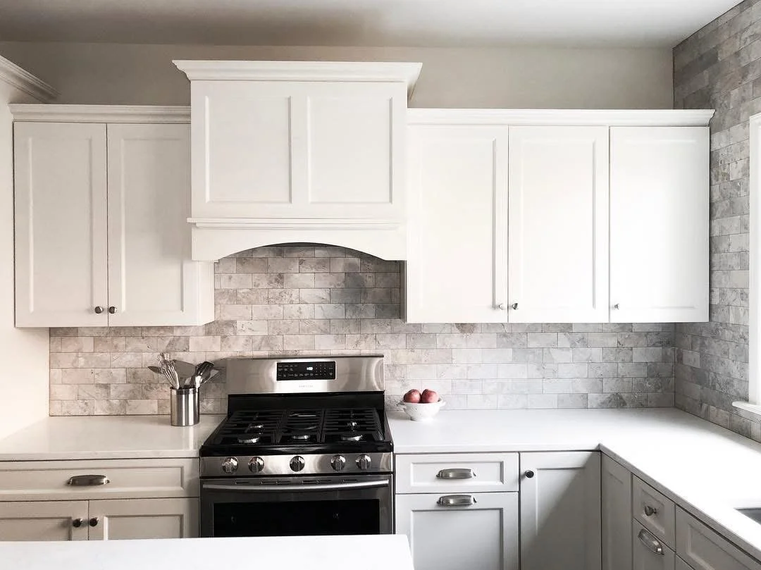 White kitchen cabinets above a stainless steel stove with a gray stone backsplash, a bowl of apples, and a container with kitchen utensils.