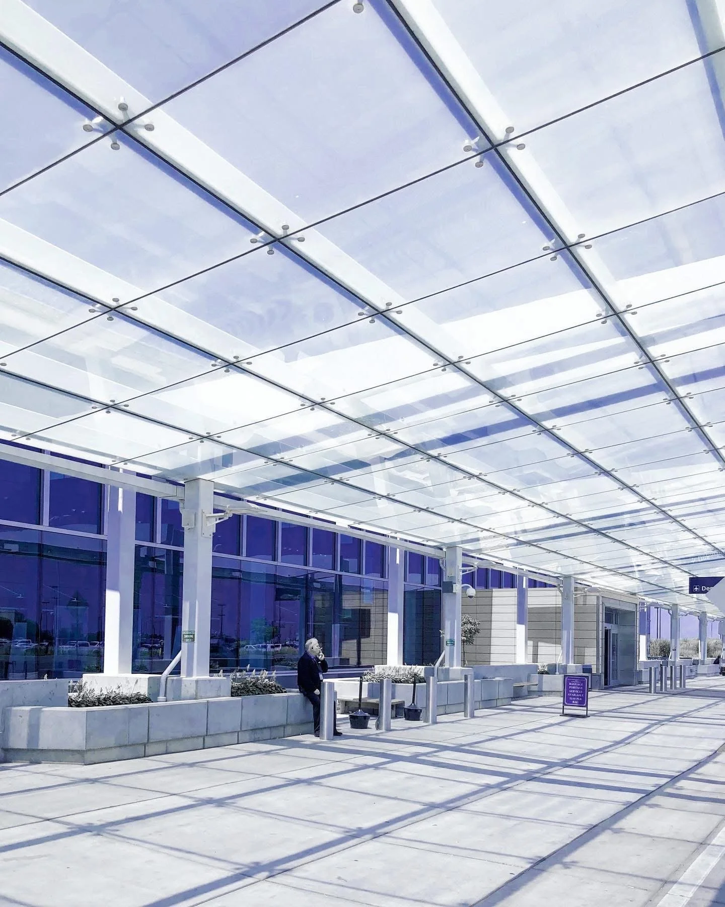 Modern airport terminal exterior with glass canopy overhead, a person sitting on a bench using a phone, and purple informational signage.
