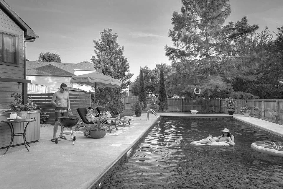 A backyard scene with a swimming pool, three people relaxing, one person grilling, and another jumping into the pool, with trees and a fence in the background.