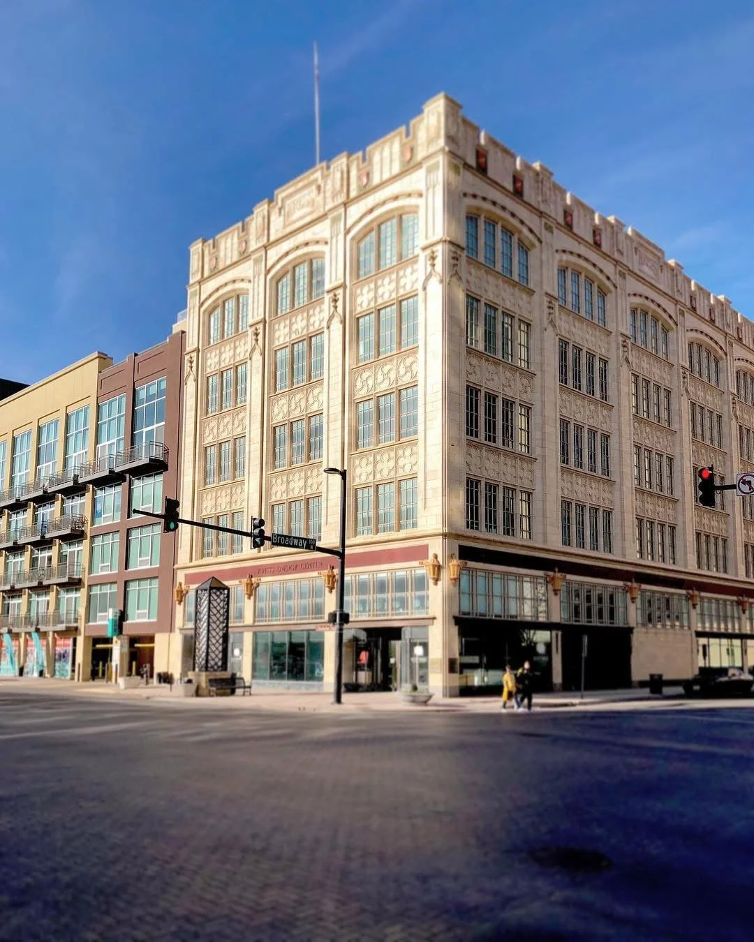 A multi-story building with ornate beige stone façade on a city street corner under a clear blue sky, with traffic lights and street signs visible.