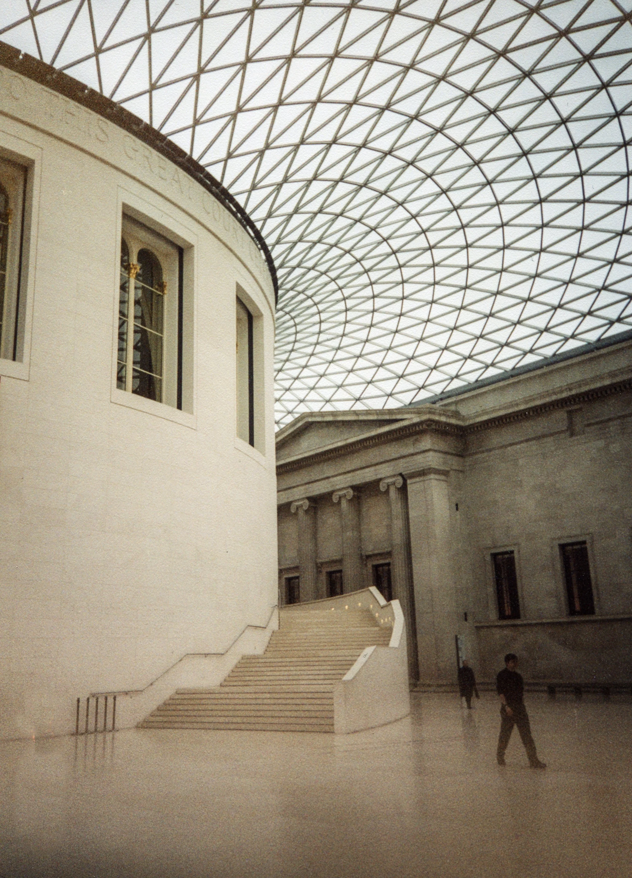 Interior view of a large historic building with modern glass and steel ceiling, stone walls, staircase, and a few people walking.