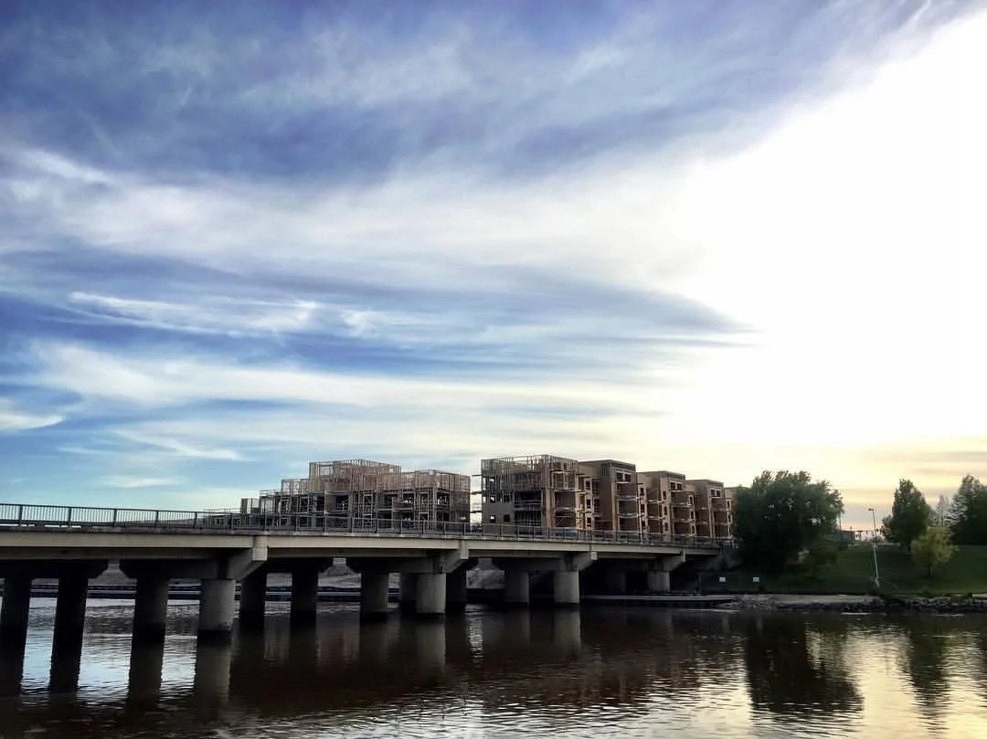 Construction site with multiple partially built buildings on a bridge over a body of water, with a partly cloudy sky and trees in the background.