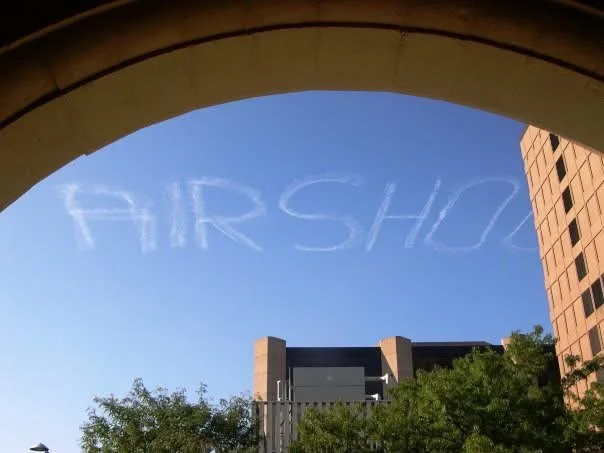 Clouds in the sky spell out the word 'AIRSHO' seen through an archway, with buildings and trees below.