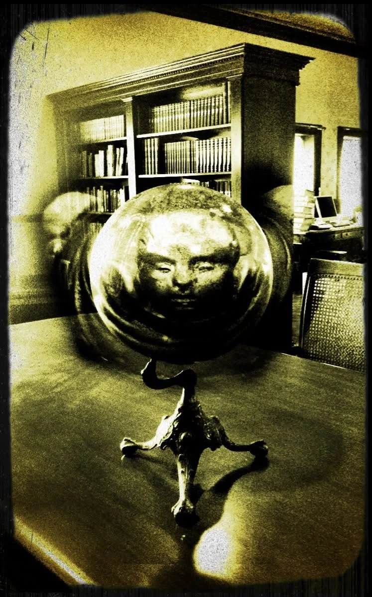 A decorative globe with a face inside the globe on a mount, placed on a table in a room with bookshelves and a computer in the background.