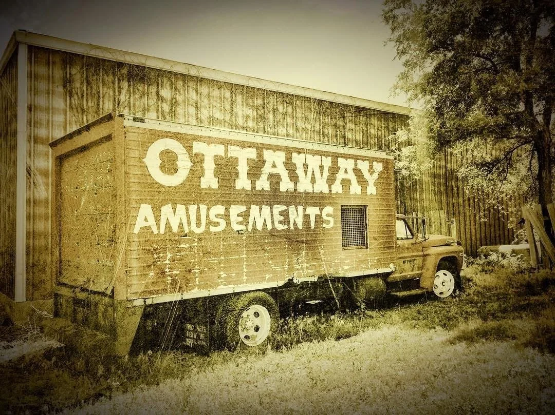 An old wooden trailer with the words 'Oklahoma Amusements' painted on the side, parked beside a corrugated metal building with trees in the background.