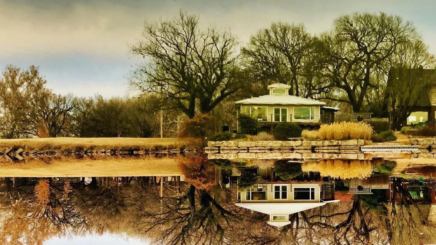 A house near a pond with its reflection visible in the water, surrounded by trees with autumn foliage.