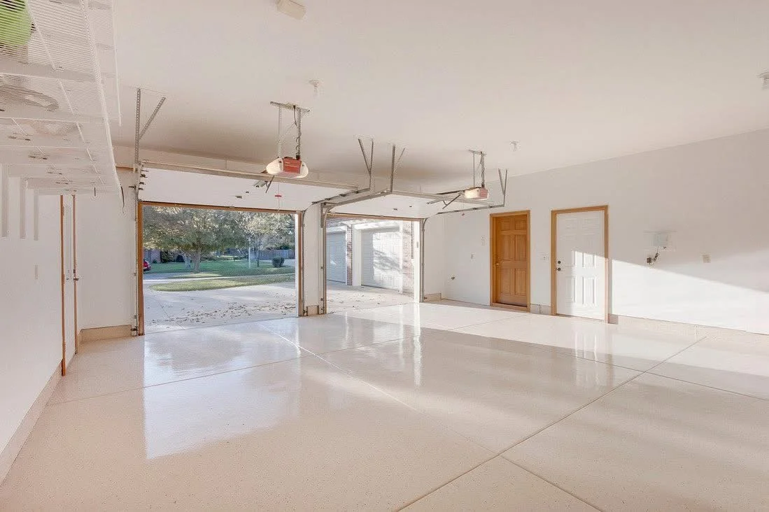 Empty garage with open doors, smooth concrete floor, and white walls, with some garage door mechanisms visible.