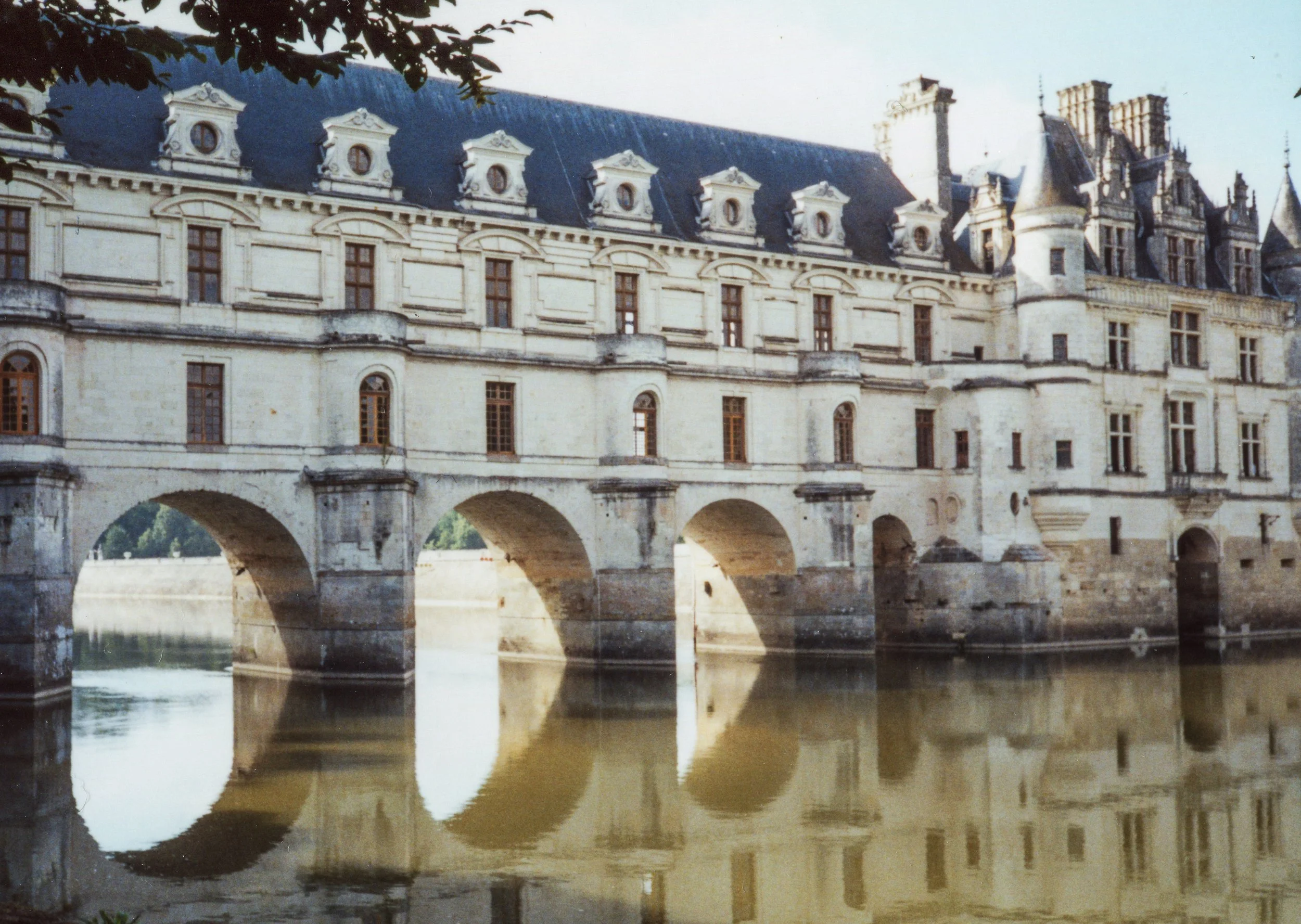 A historic chateau with multiple windows and towers, reflected in a calm body of water.