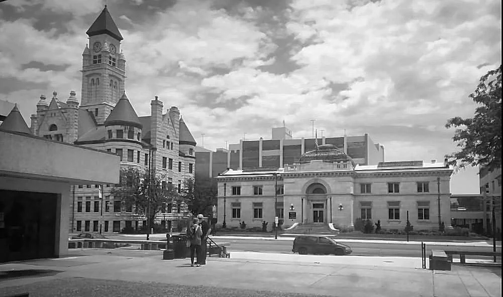 A black and white photo of a historic courthouse with classical architecture, surrounded by other buildings including a castle-like structure with turrets. People are standing near the sidewalk in front.