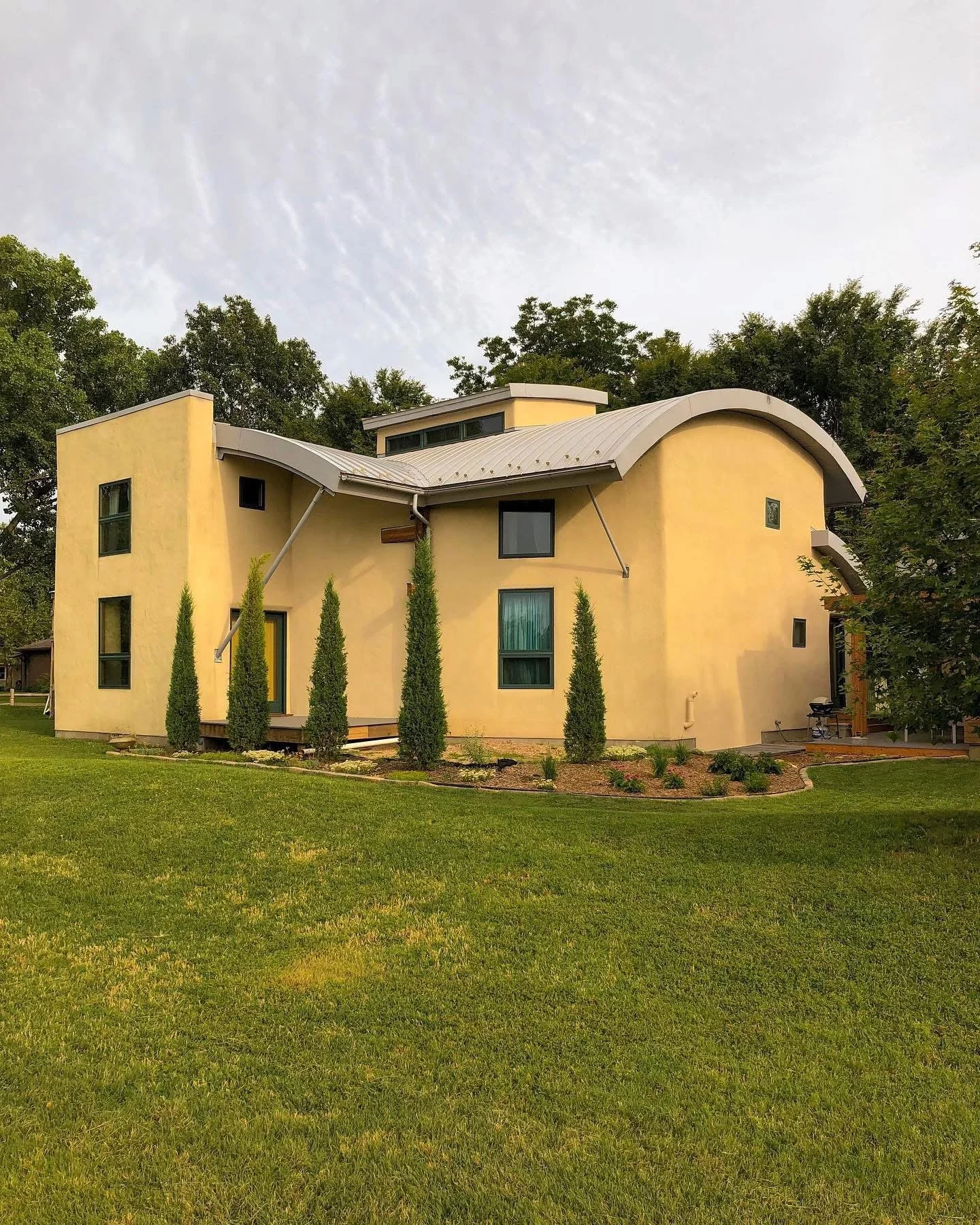 A modern, yellow, two-story house with a curved metal roof and tall, narrow trees in the front yard.