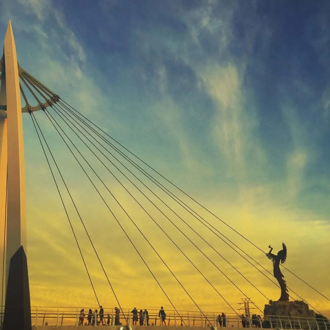 People walking on a bridge with a large suspension structure and a statue of a woman holding a torch against a sunset sky.