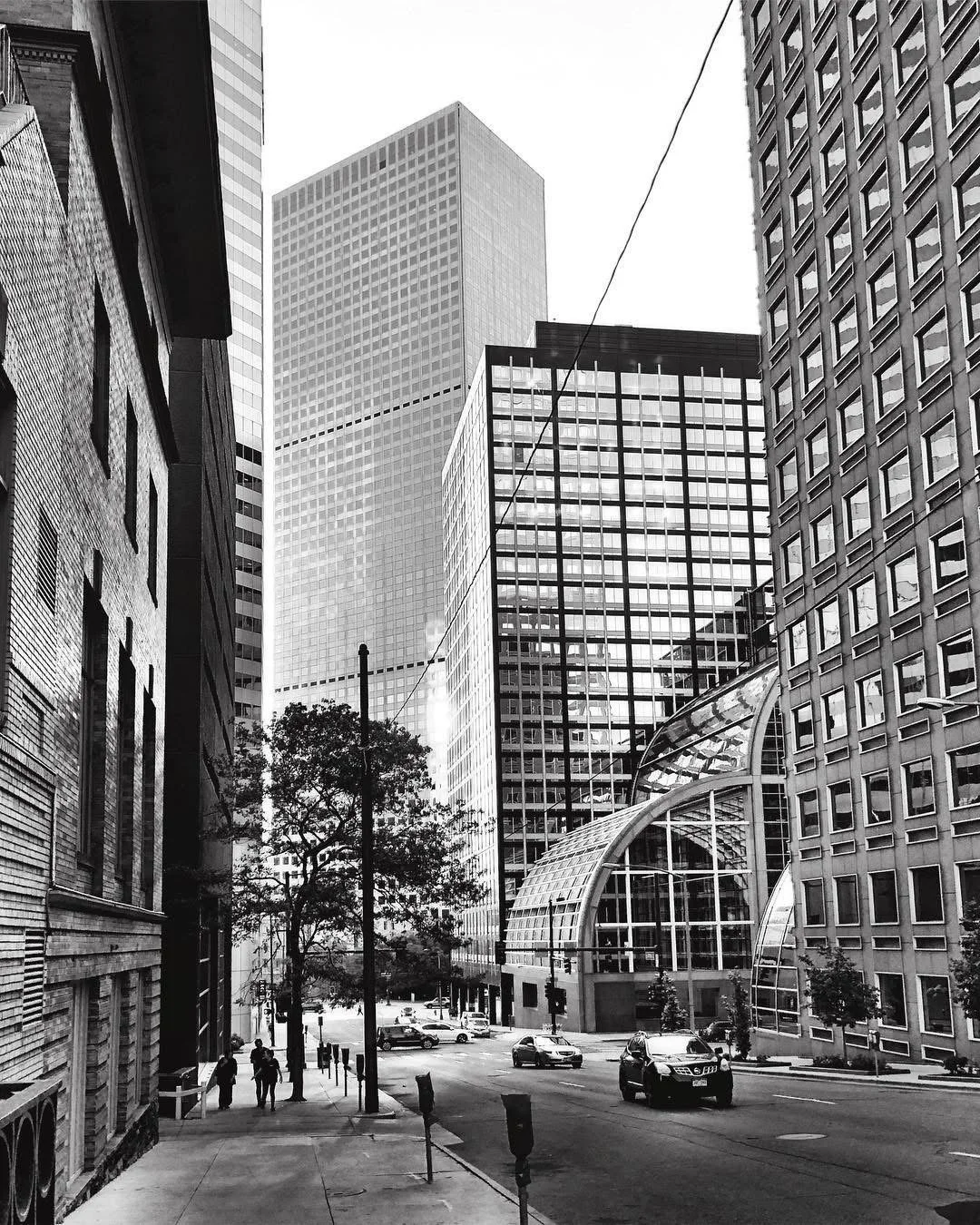 Black and white photo of tall modern city buildings with glass facades, a street with cars, pedestrians, trees, and a distinctive curved glass roof entrance.