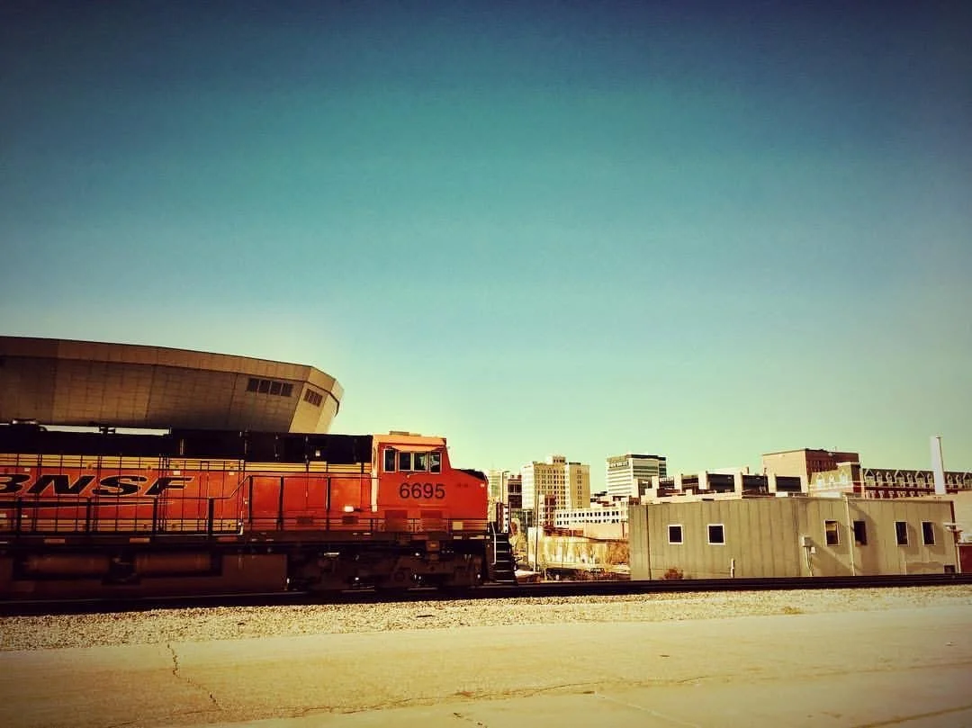 A red train passing through an urban area with buildings in the background and a clear blue sky.