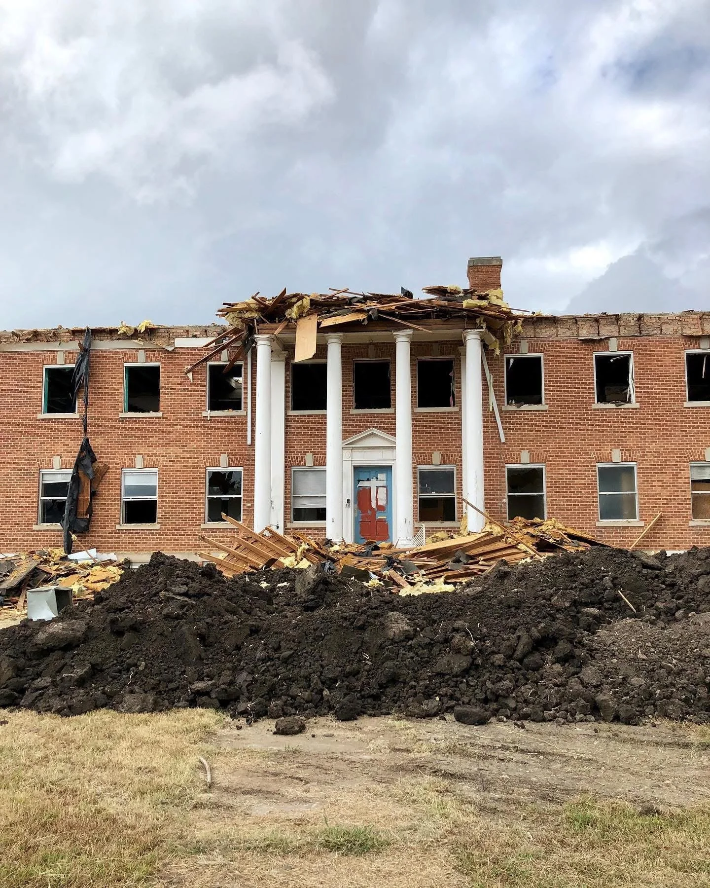 A large brick building with extensive damage, missing windows, and a partially collapsed roof. Debris and dirt are piled in front of the building, with dark clouds overhead.