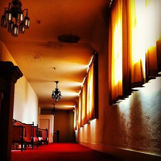 Long hallway with yellow curtains along windows, vintage chandelier lighting fixtures, wooden furniture, and red carpet.