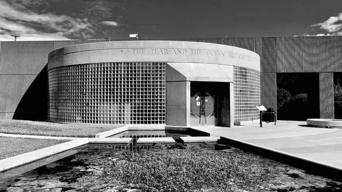 Black and white photo of a modern building with a curved glass block facade and a sign that reads, 'The tear and the ocean are sisters.'
