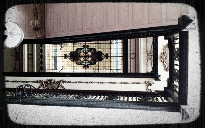 Interior view of a building with ornate details, a staircase, and a decorative ceiling with stained glass window, seen from below.