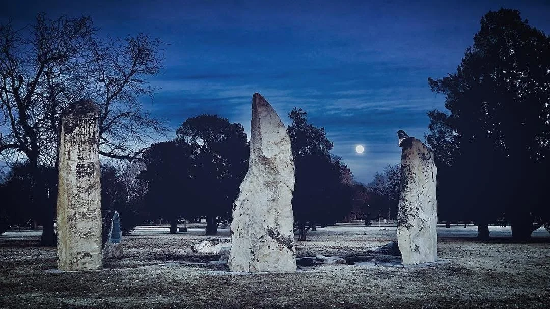 Three large white standing stones in a park at night, under a moonlit sky with trees in the background.