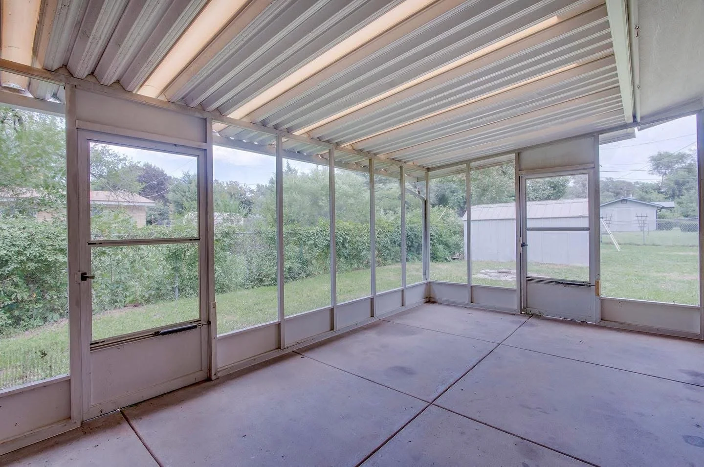 Unscreened porch with screened walls, concrete floor, and metal roof, overlooking a backyard with grass and trees.