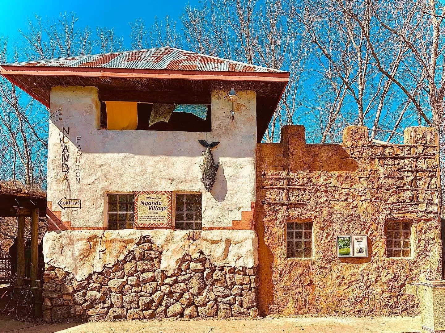 Facade of Nganda Village building made of stones and stucco with a fish mounted on the wall, signage indicating it is Nganda Village established in 2004, and a sign directing to gorillas. The structure has a metal roof and windows, with bare trees in