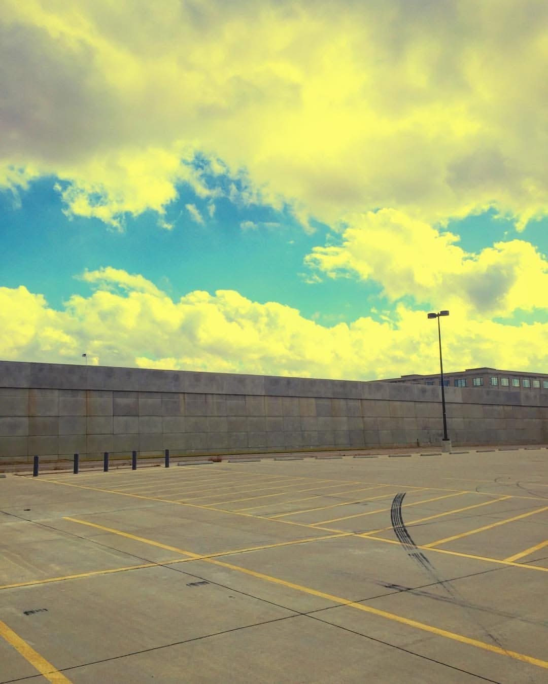 Empty parking lot with yellow lines, a stone wall, a street lamp, and a cloudy sky with yellow-tinted clouds and blue patches.
