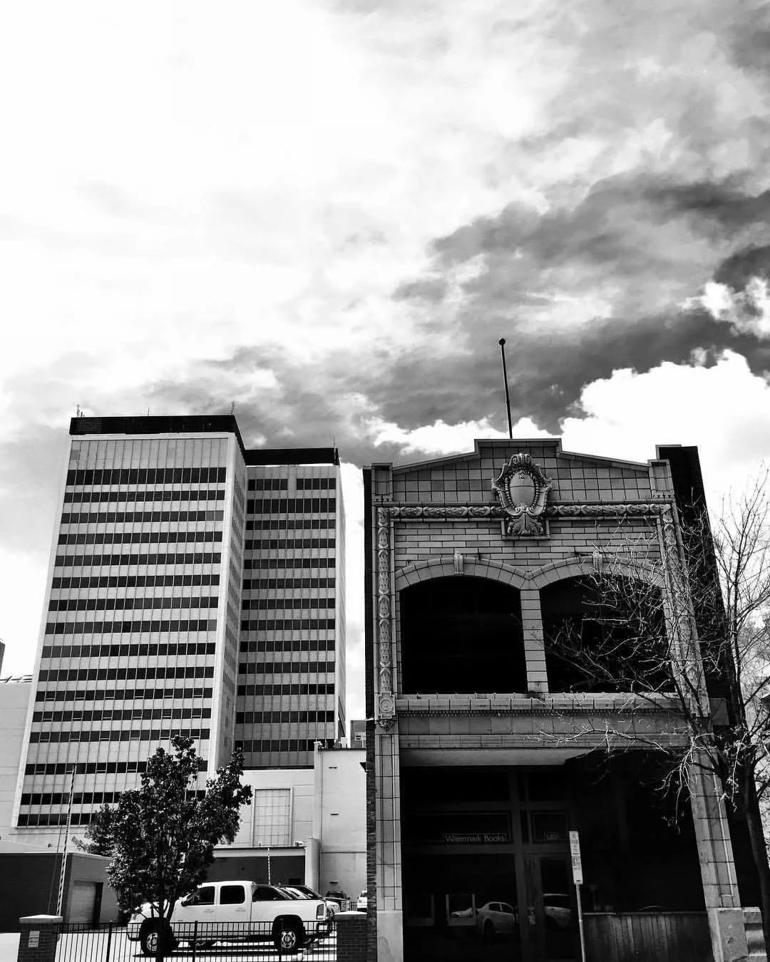 Black and white photo of tall modern office buildings, an old ornate building, a parked truck, a small tree, and cloudy sky