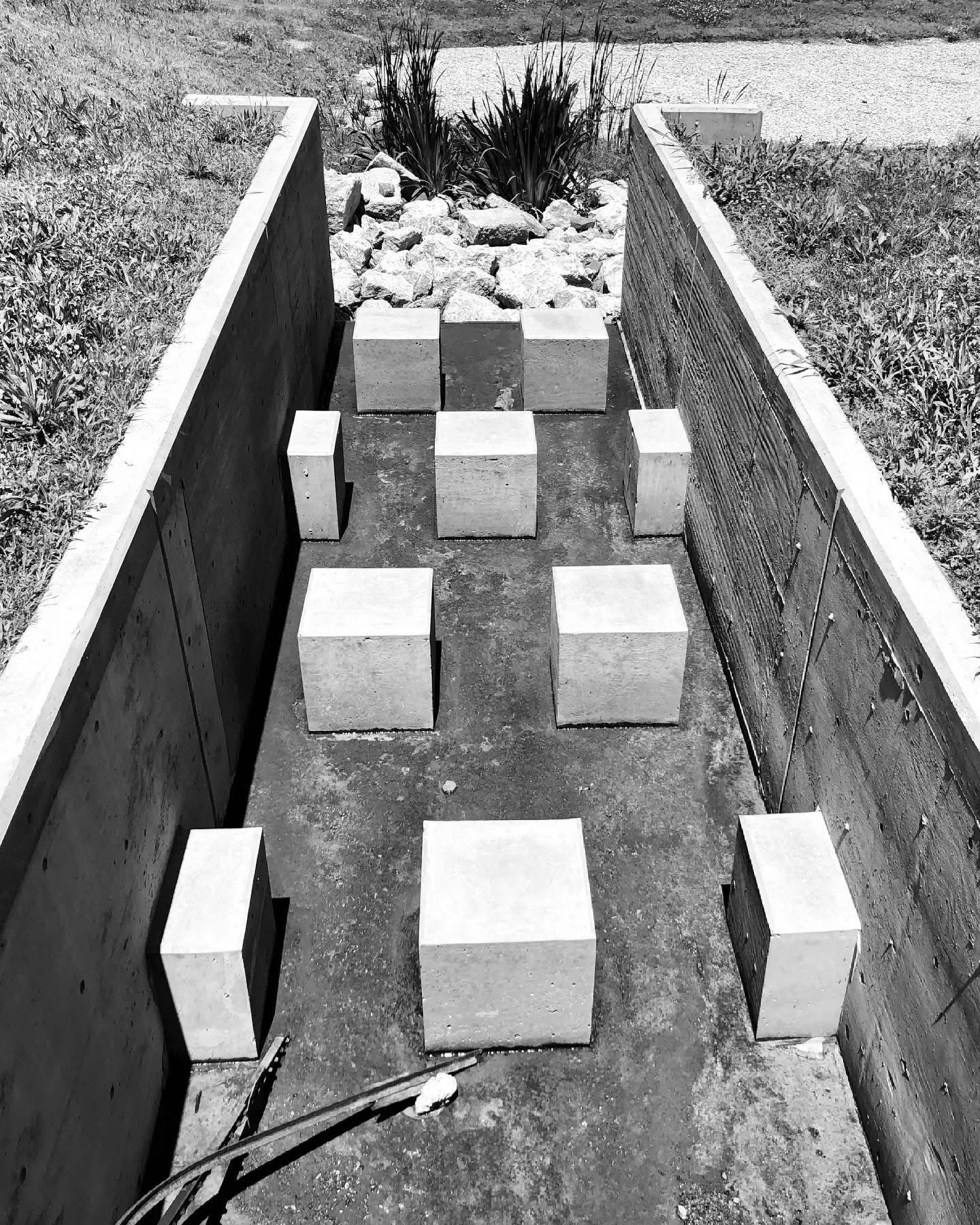 A construction site with a long, narrow trench lined with concrete walls. Inside the trench, several concrete blocks are arranged, and at the end, there are rocks and plants beyond the trench.