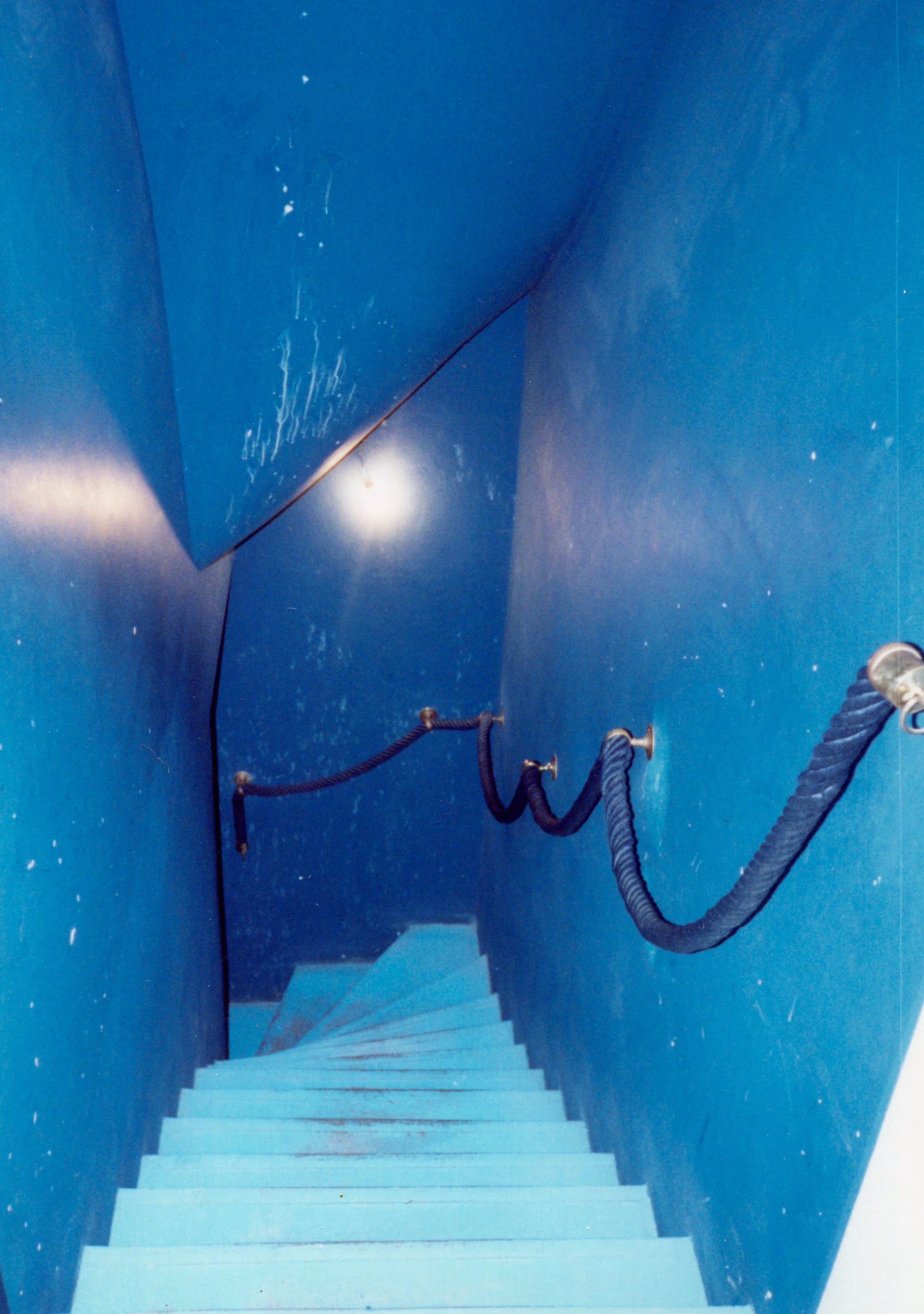 Blue staircase with a black rope handrail on the right side, walls with visible scratches and scuffs, and a ceiling with a spotlight or light source.