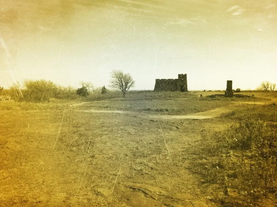 A barren landscape with a dirt path leading to an old, abandoned castle on a hill. There are a few leafless trees and sparse bushes, with a yellowish sky.