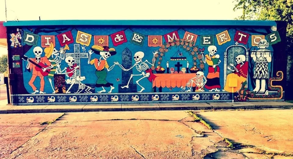 Colorful mural celebrating Día de los Muertos with skeletons participating in festivities, including playing guitar, dancing, and offering food at an altar, with festive banners above.