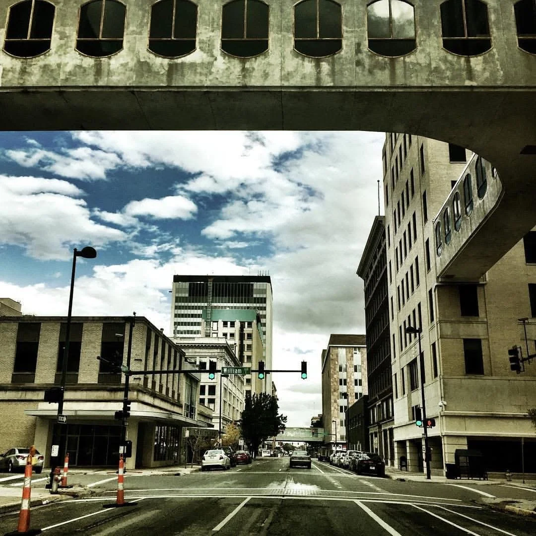 City street scene with tall buildings, traffic lights, cars parked along the curb, and a partly cloudy sky overhead.